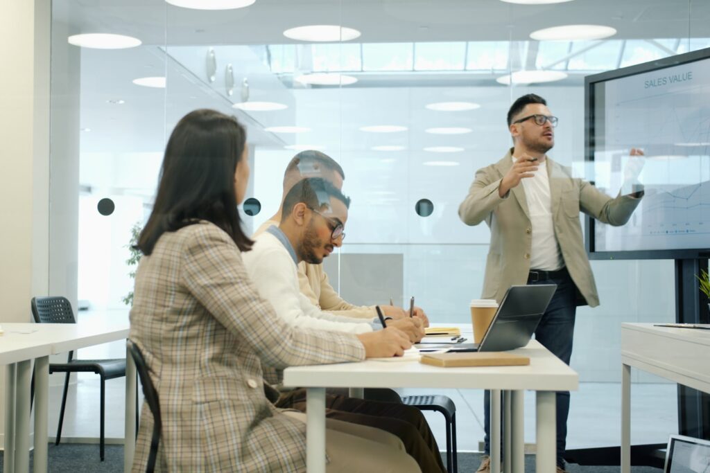 Group of people men and women are listening to boss during meeting in boardroom, middle-aged man is using digital screen for presentation in office.